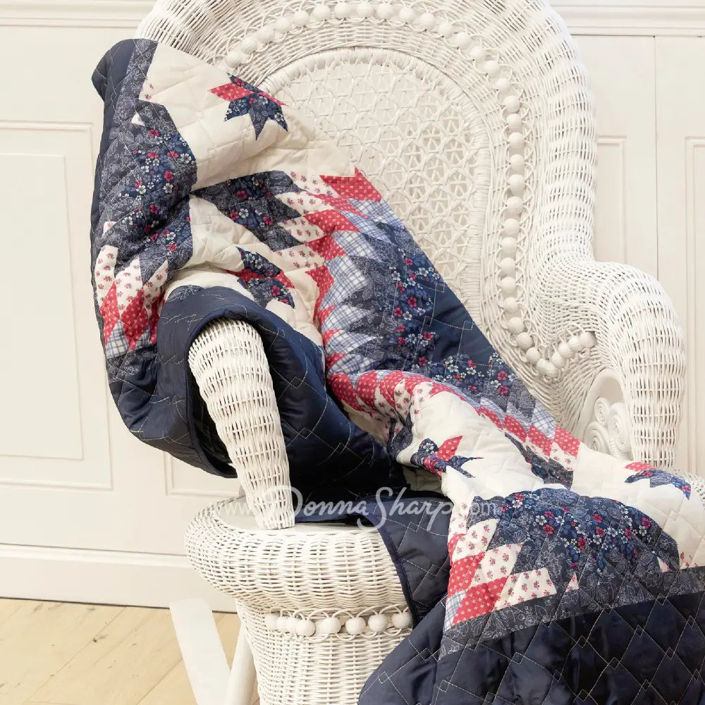 Donna Sharp Bluefield Star reversible throw blanket draped on white wicker chair showing a floral star pattern with navy, country blue, periwinkle, pale ivory, rose, and raspberry, with a solid navy reverse side