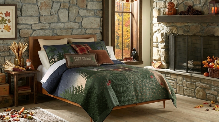 A rustic bedroom scene with a bed dressed in a forest-patterned green quilt and matching pillows, set in a stone-walled room with a fireplace, window, and autumn decor.