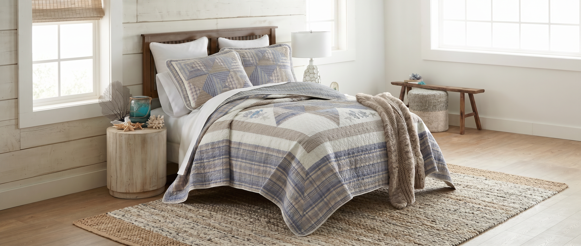 A rustic-style bed in a bright, shiplap bedroom, styled with a blue, beige, and white patchwork quilt, matching pillow shams, a wooden headboard, and a woven rug.